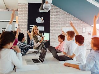 Schüler*innen sitzen mit Lehrerin am Tisch mit Laptops vor sich.